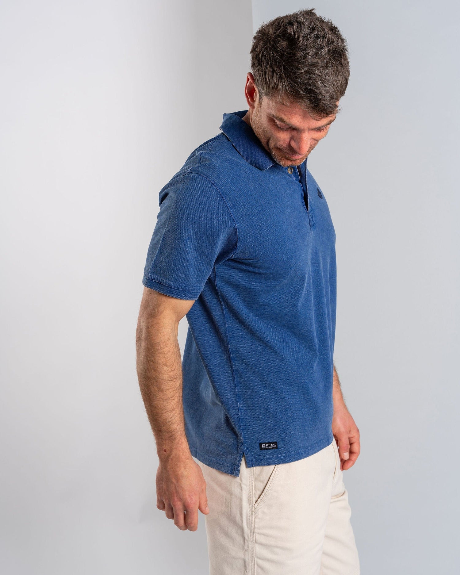 A man models the blue Saltrock Seb men's short sleeve polo made of 100% cotton, paired with white pants, against a plain background.