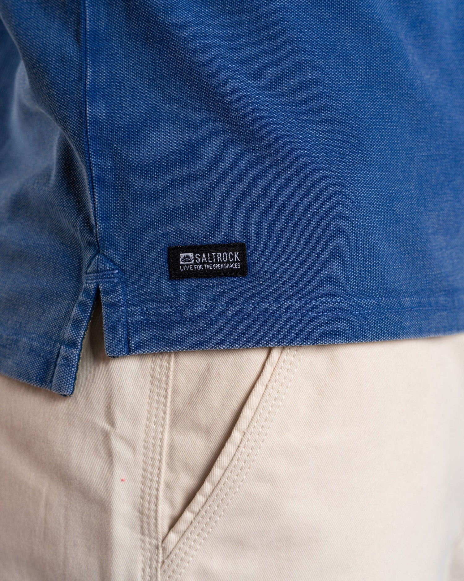 A person is wearing the Seb Men's Short Sleeve Polo in blue by Saltrock, paired with beige pants. The 100% cotton shirt features a small black label reading "Saltrock Live for the weekends.
