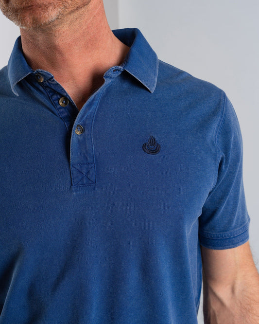 A man poses against a light background in the Saltrock "Seb" blue polo, crafted from 100% cotton, featuring a small embroidered flame logo for added style.