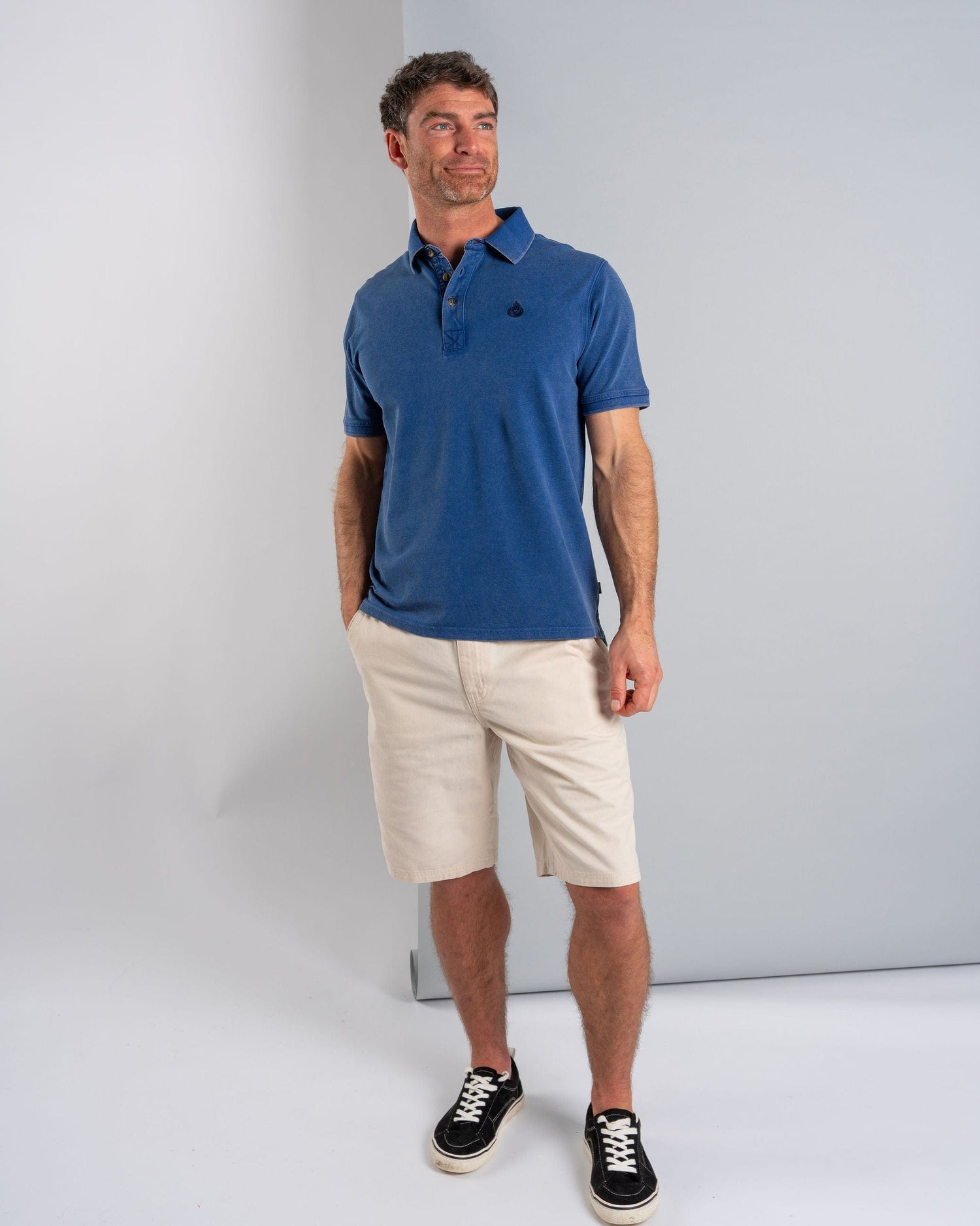 Wearing the Seb Men's Short Sleeve Polo in blue by Saltrock, a man pairs it with beige shorts and black sneakers, standing against a gray background while gazing thoughtfully to the side.