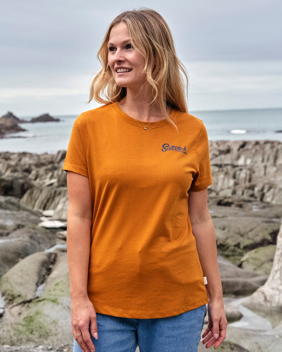 A woman wearing a lightweight fabric yellow t-shirt from the Saltrock brand, standing on a rocky beach.