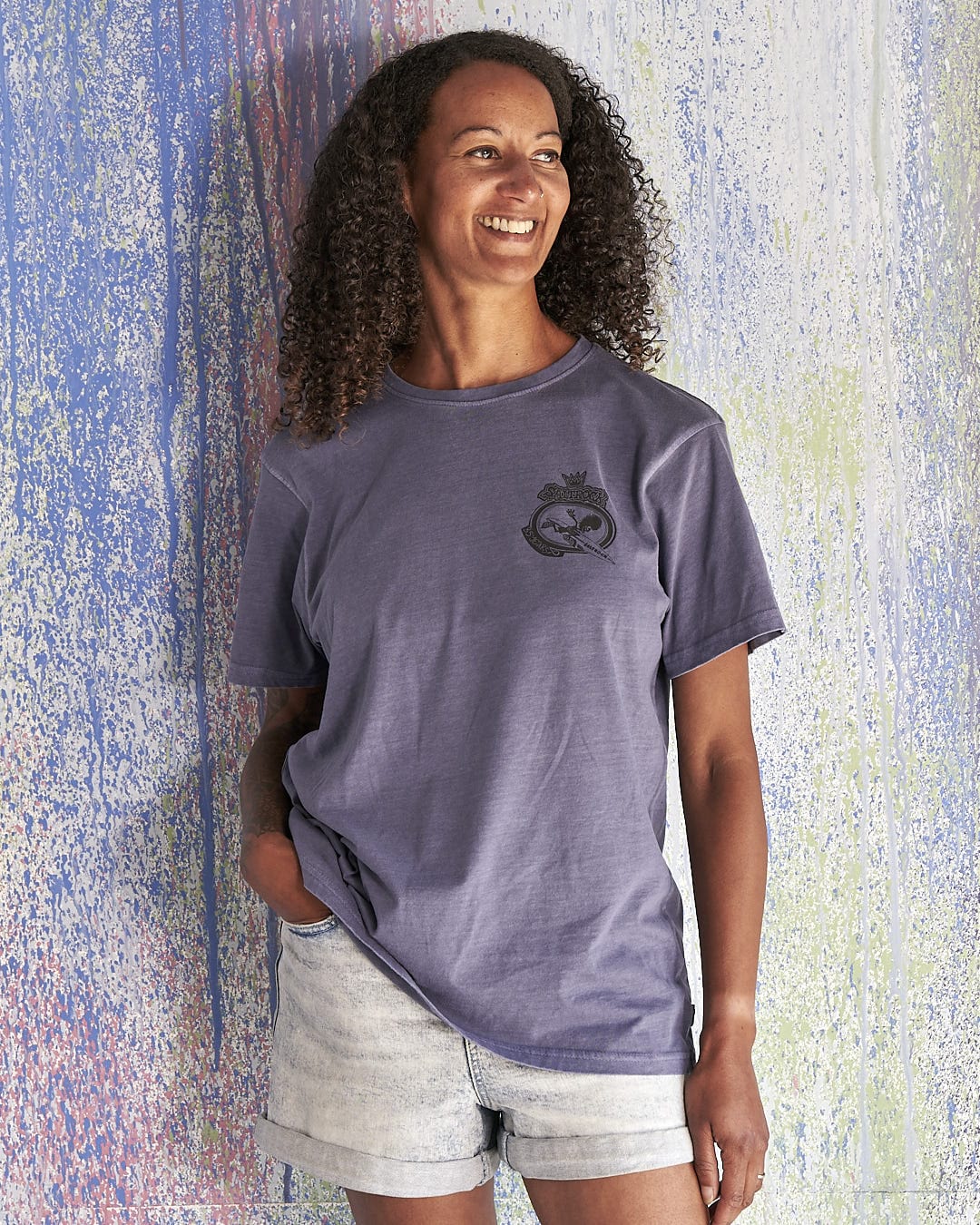 A woman wearing a Tribal Saltrock - Limited Edition 35 Years T-Shirt and shorts is leaning against a wall.