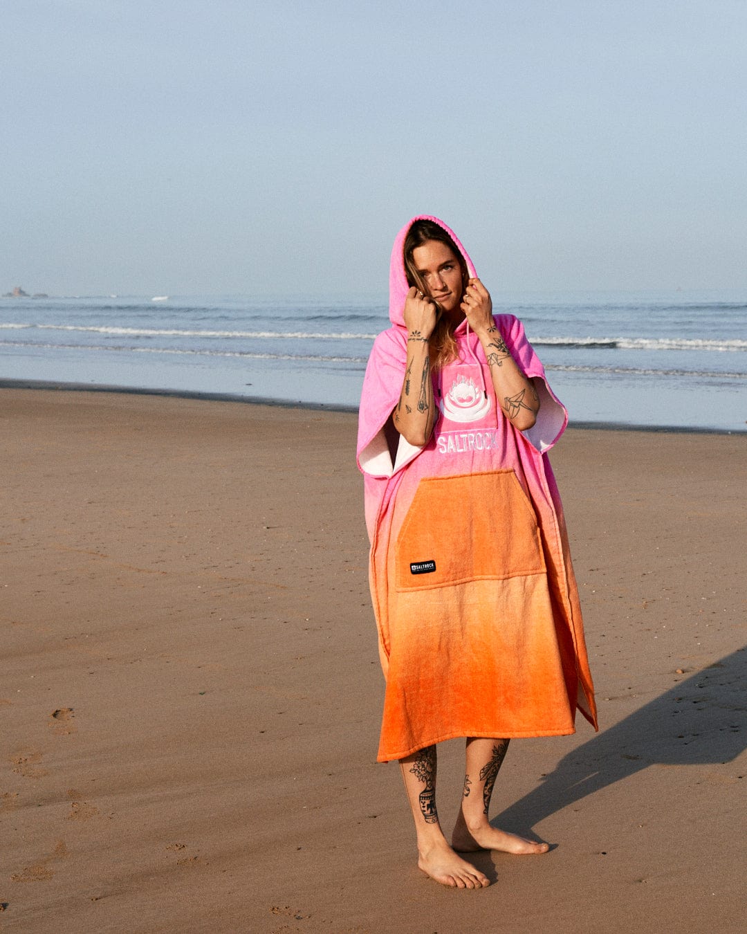 A person in a pink hoodie and a Saltrock Tropic Dip - Changing Towel - Pink/Orange stands barefoot on a sandy beach, touching their hood, with waves in the background.