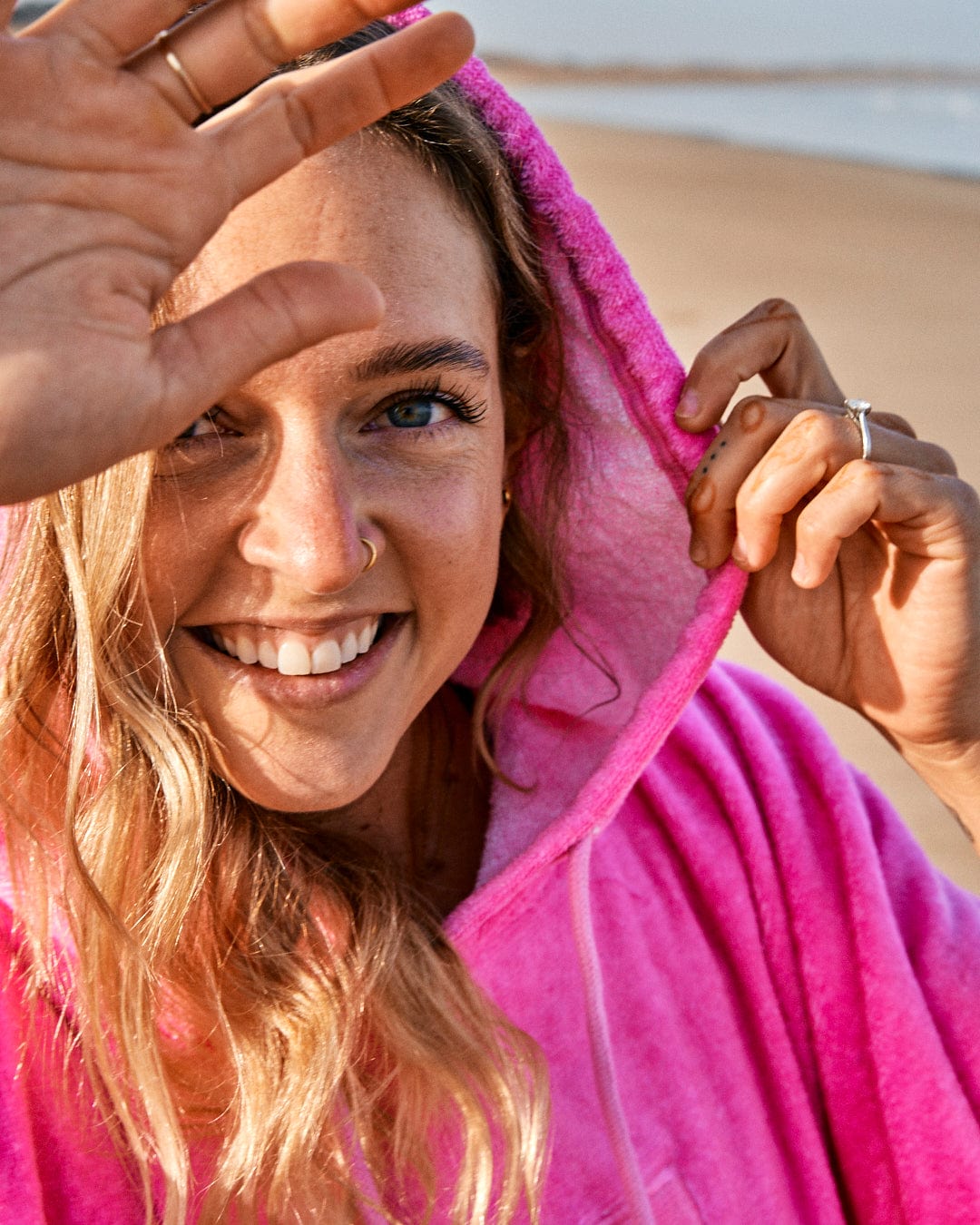 Woman in a pink, fast-drying Tropic Dip Changing Towel - Pink/Orange hoodie smiling at the camera, shielding her eyes from the sun with her hand, at the beach. (Saltrock)