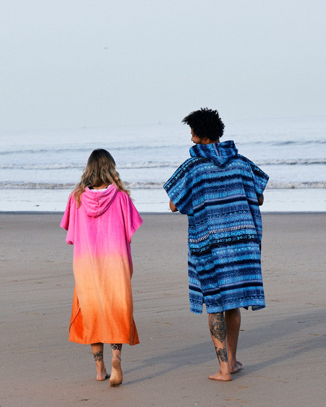 Two people wrapped in colorful, ultra-absorbent Saltrock Tropic Dip - Changing Towels in Pink/Orange walking on a beach, viewed from behind, with clear skies in the background.