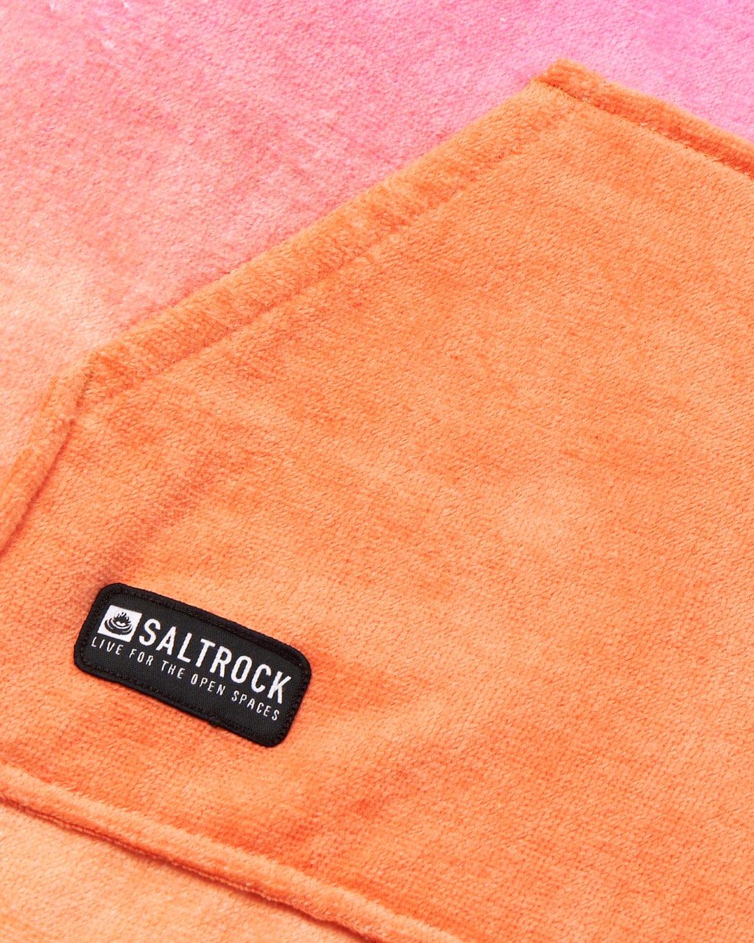 Close-up of an ultra absorbent Tropic Dip - Changing Towel - Pink/Orange with the Saltrock logo, set against a pink and red watercolor background.