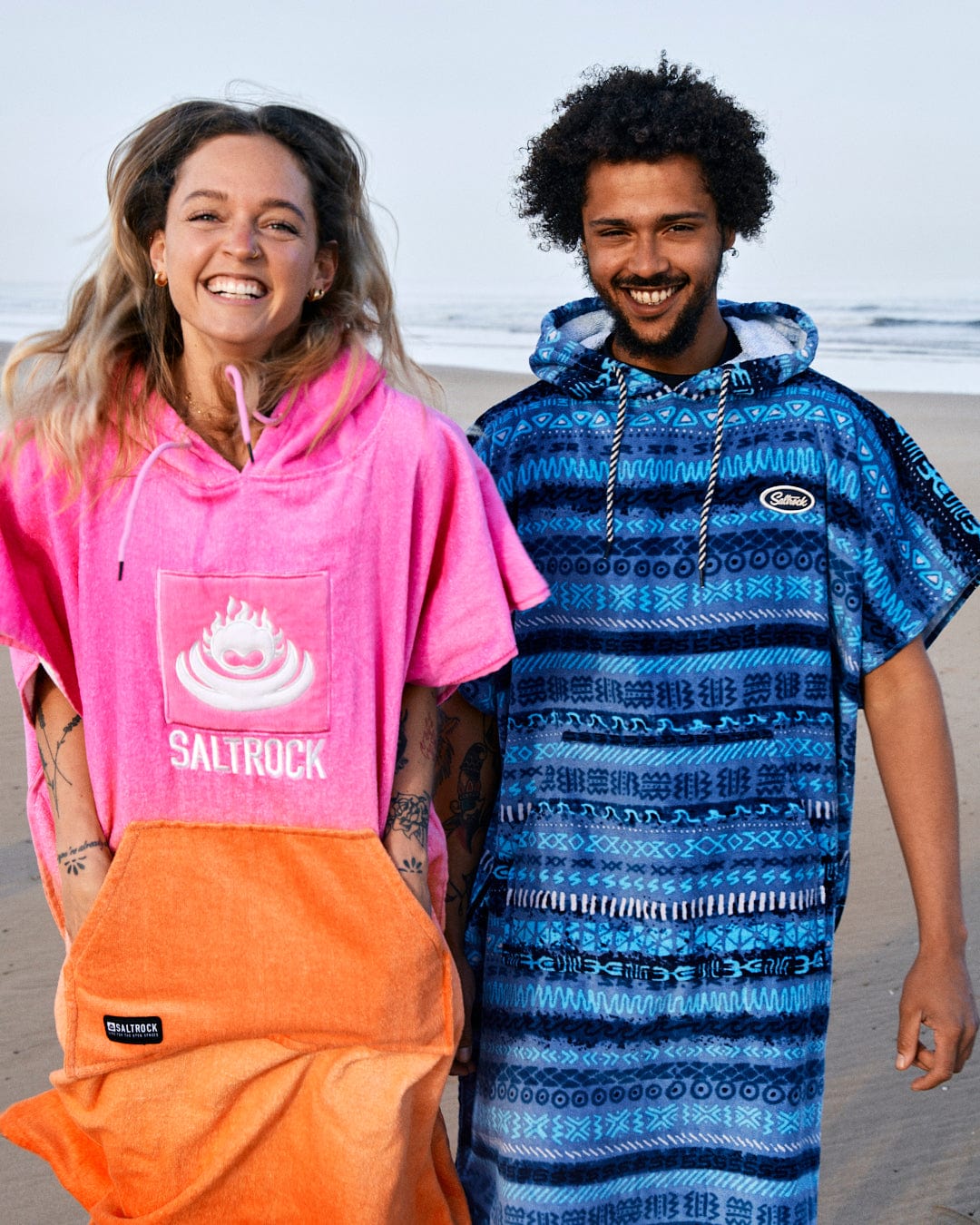 Two young adults smiling on a beach, one wearing a Saltrock bright pink Tropic Dip Changing Towel and the other in a Saltrock blue patterned hoodie made of 100% cotton, both casually dressed with visible tattoos.