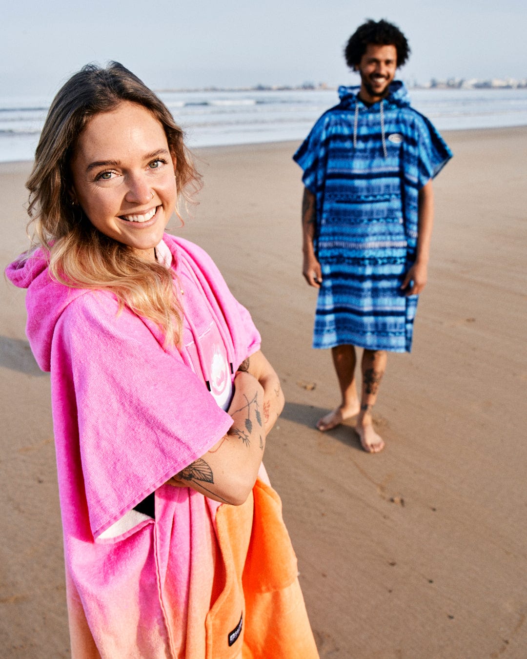 A woman in a Saltrock Tropic Dip - Changing Towel - Pink/Orange poncho smiling at the camera on a beach with a man in a blue poncho standing behind her, sand and waves in the background.