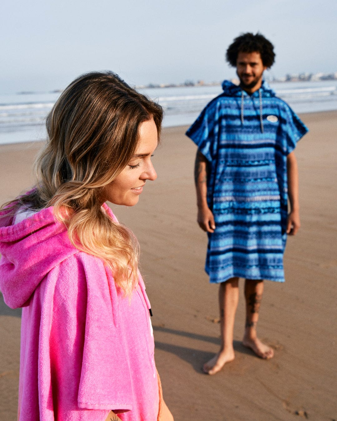 A woman in a pink Tropic Dip changing towel from Saltrock and a man in a blue poncho stand on a beach, with the woman looking to the side and the man towards her in the background.
