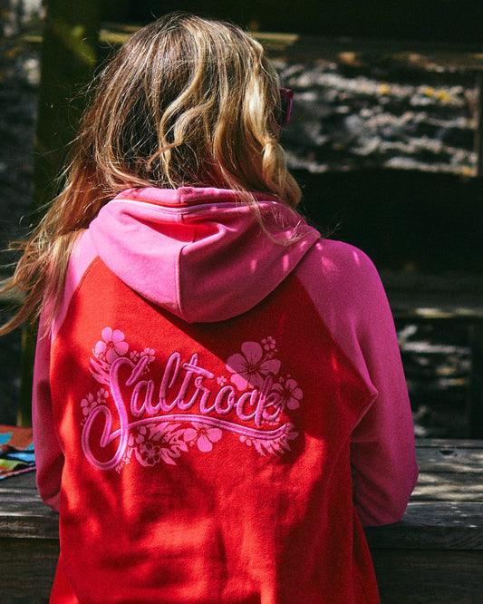 A person with long hair is sitting outdoors, wearing a Saltrock Tropic Women's Zip Hoodie in red with a floral design on the back, featuring Saltrock branding.