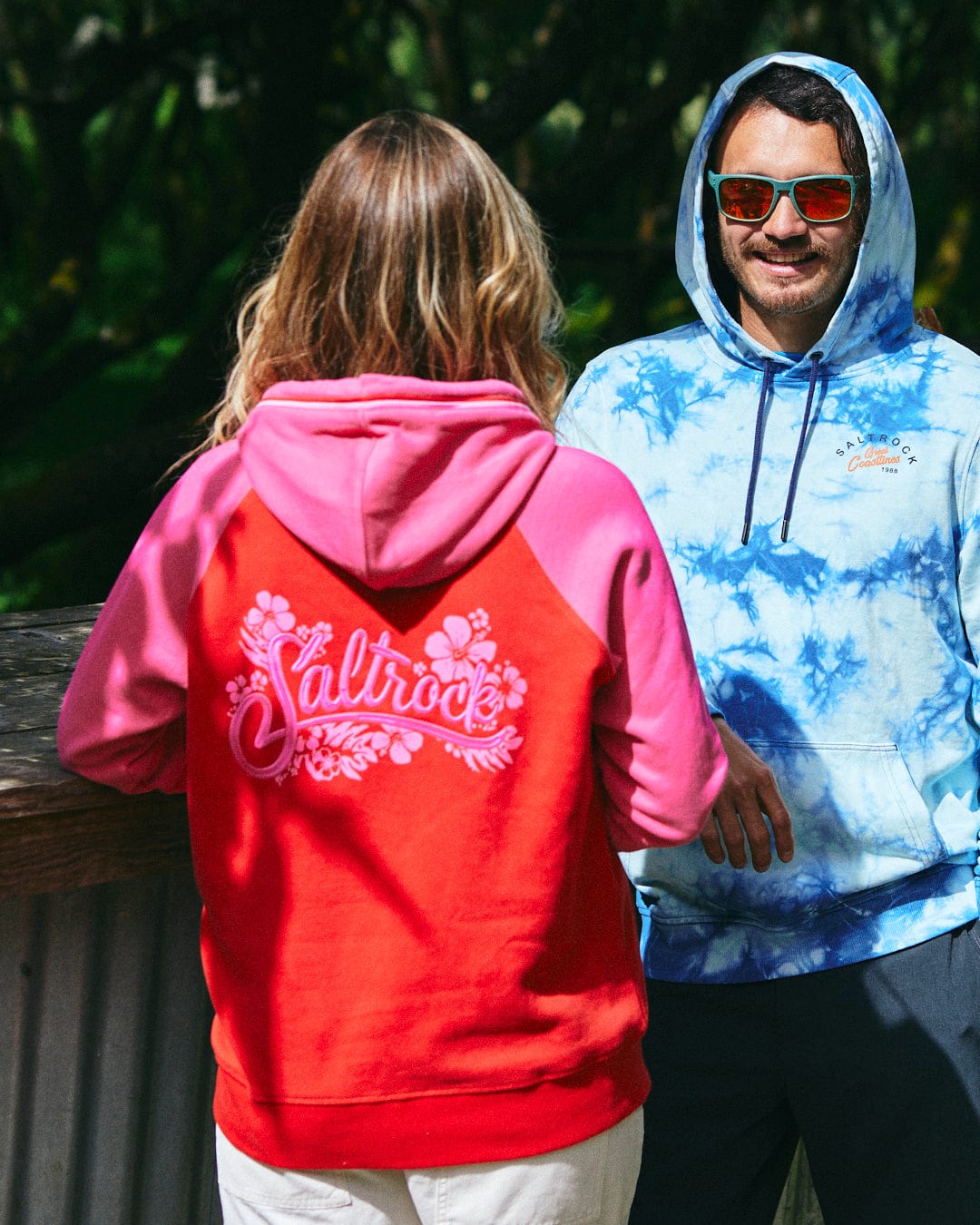 Two people are standing outdoors, one wearing the Saltrock Tropic women's zip hoodie in red with an embroidered floral design on the back, and the other in a blue tie-dye cotton hoodie with sunglasses.