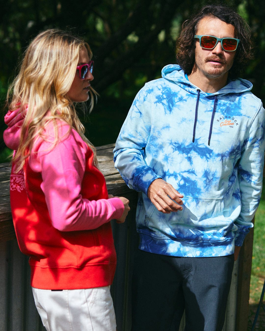 Two individuals wearing colorful sunglasses and Saltrock-branded hoodies—one in the Tropic Women's Zip Hoodie in Red and the other in a blue tie-dye hoodie—stand outdoors near a wooden structure with greenery in the background.