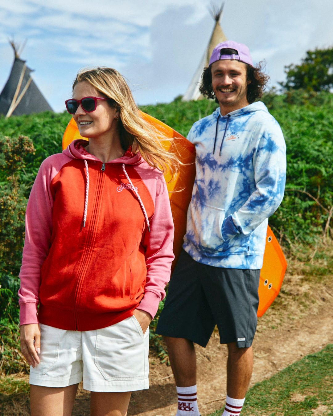 Two people stand outdoors; one wears the Saltrock Tropic - Womens Zip Hoodie in red with white shorts, and the other a blue tie-dye hoodie with gray shorts. They are smiling, with one holding a bright orange surfboard.