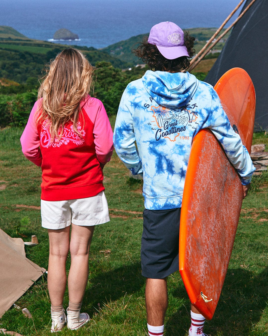 Two people stand outdoors facing away. The person on the left wears a red Saltrock Tropic Women's Zip Hoodie and white shorts; the one on the right sports a blue tie-dye hoodie with Saltrock branding, black shorts, a purple cap, and holds an orange surfboard.