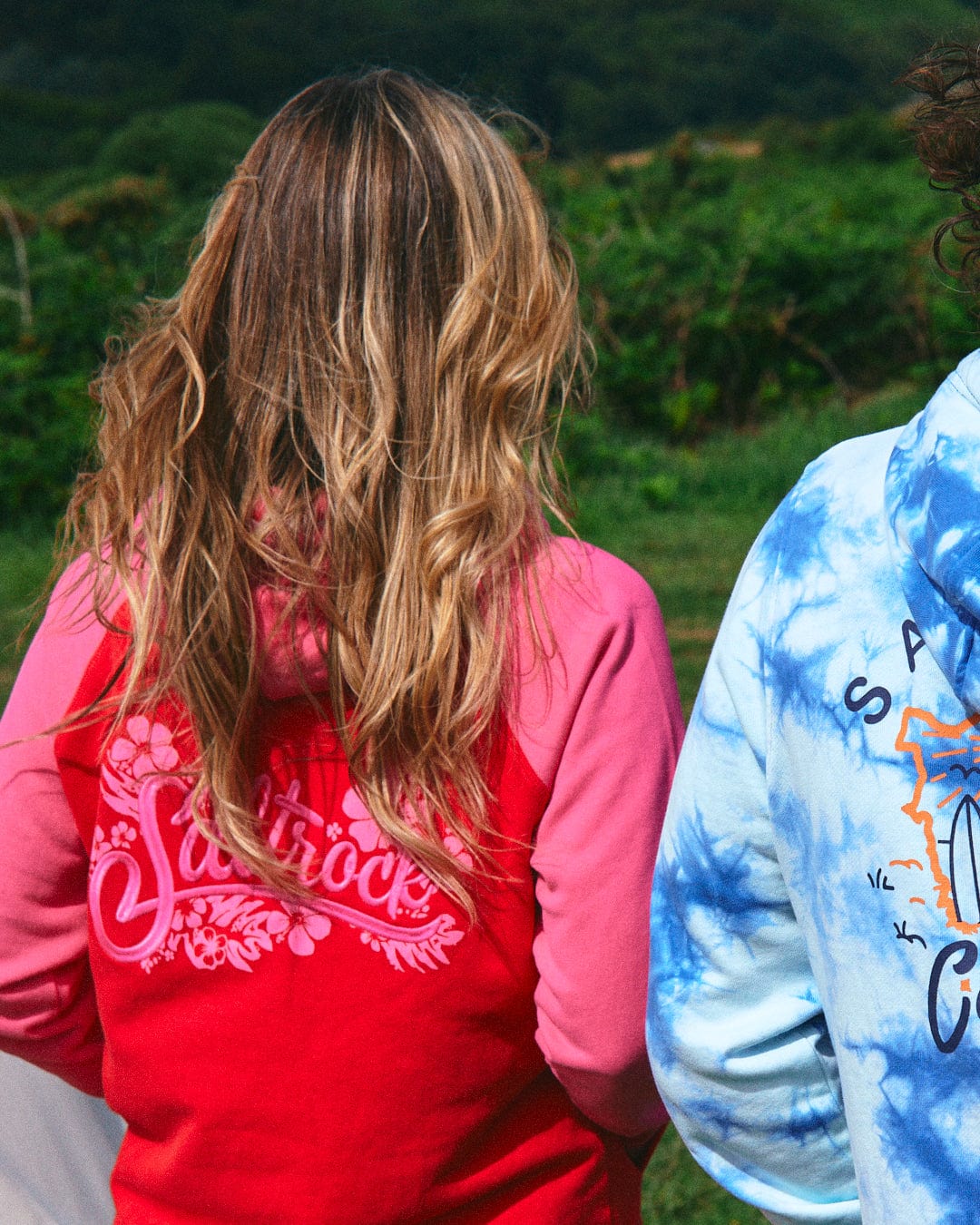 Two individuals outdoors with their backs facing the camera—one in the Tropic Women's Zip Hoodie in red from Saltrock, and the other in a blue tie-dye hoodie. Both hoodies feature subtle Saltrock branding and are made of soft cotton, perfectly blending style and comfort in a lush, green natural setting.
