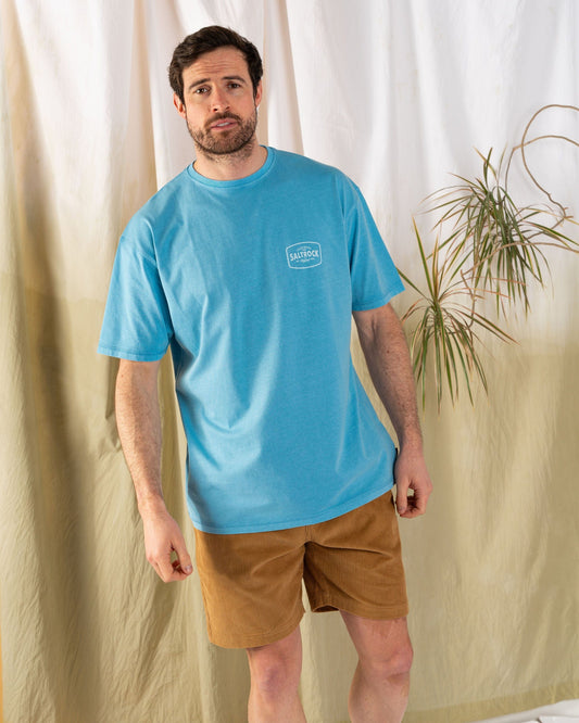 A man in an oversized, blue Saltrock Jungle Wanderer T-shirt and tan shorts stands in a room with light-colored drapes and a potted plant in the background.
