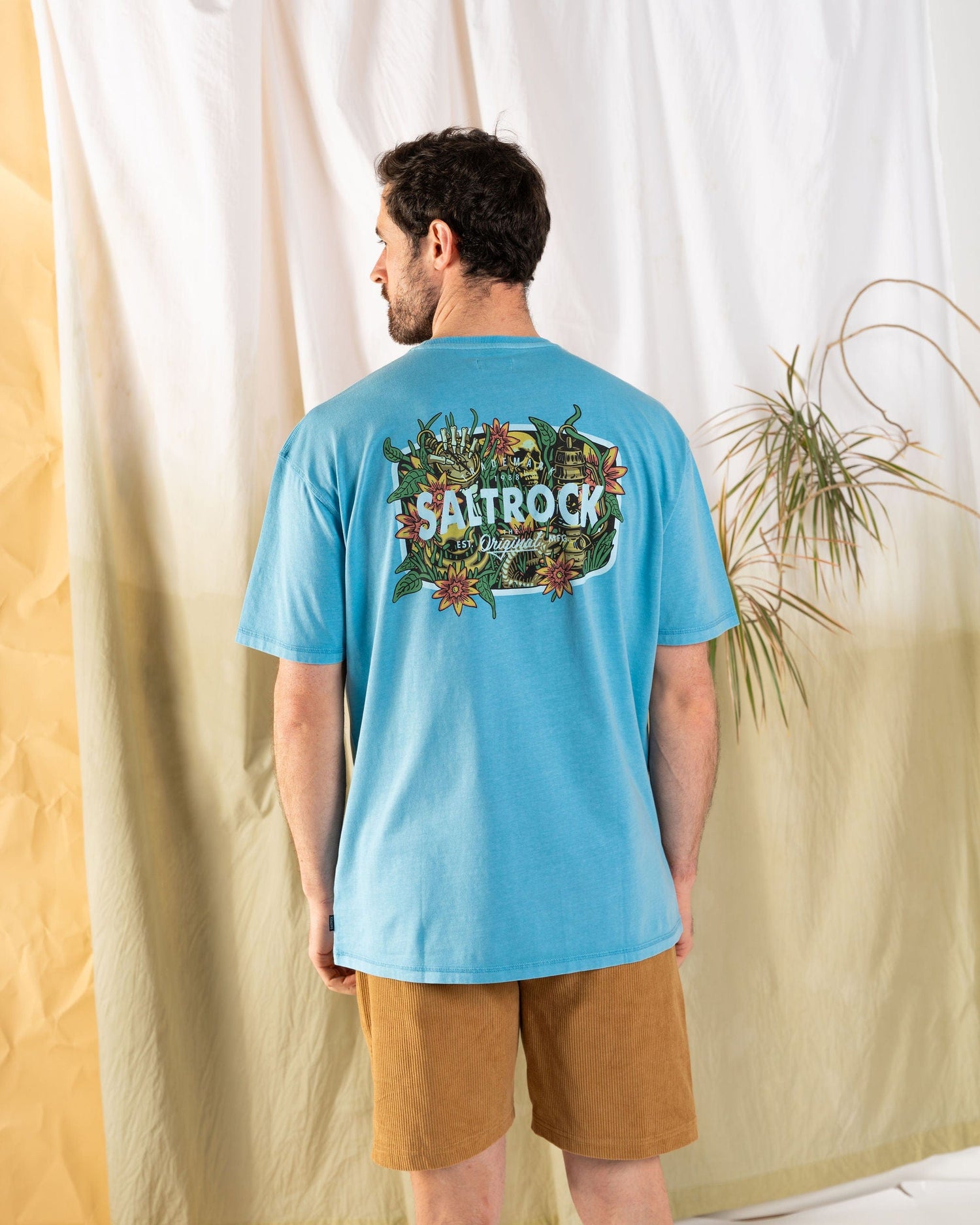 A man wearing a blue Saltrock Jungle Wanderer Men's Oversized T-Shirt and brown shorts stands facing away from the camera, against a light backdrop with a plant.