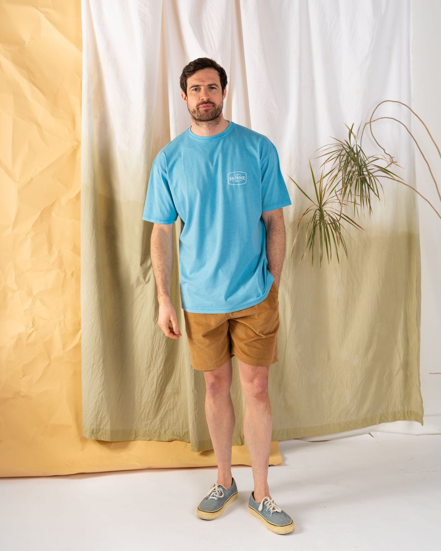 A man stands on a white floor with a beige and green fabric backdrop, wearing the Saltrock Jungle Wanderer Men's Oversized T-Shirt in blue, paired with brown shorts and gray sneakers. A plant subtly complements the scene.