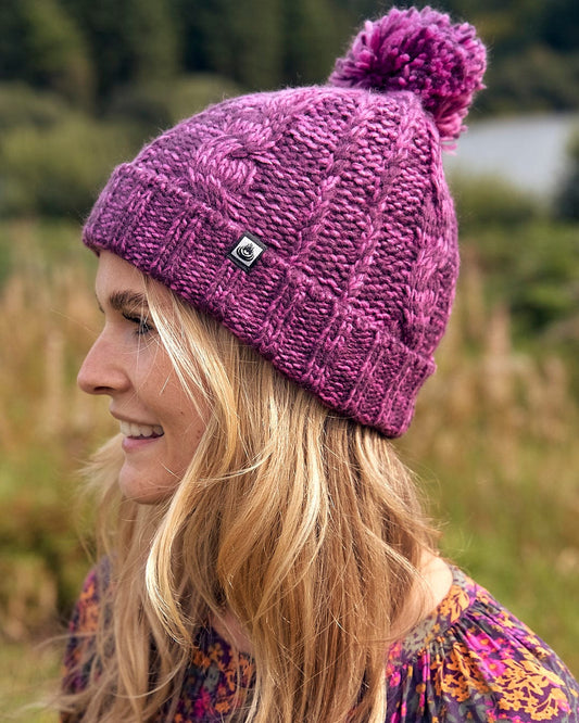 A woman wearing a Twist - Cabled Beanie - Dark Pink with a pom pom made by Saltrock.