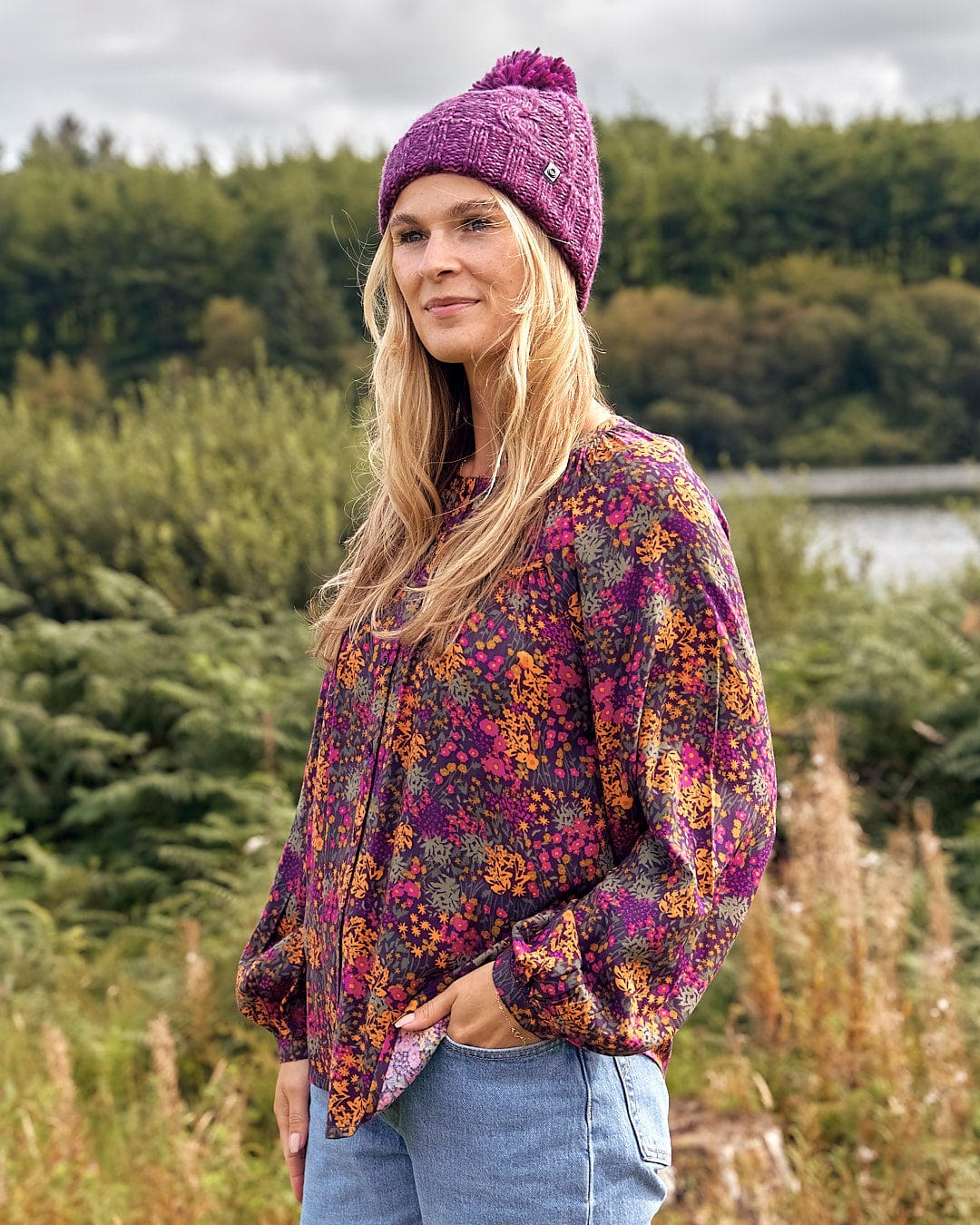 A woman wearing a Saltrock Twist - Cabled Beanie - Dark Pink and jeans standing by a lake.