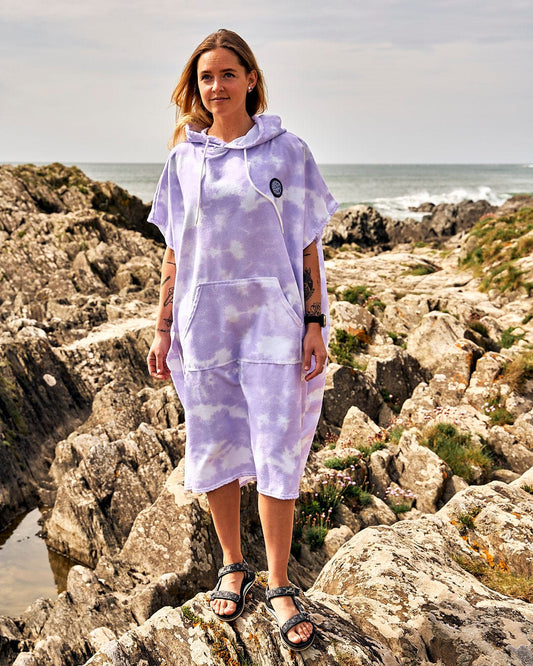 A woman wearing a Saltrock Ubud - Womens Tie Dye Changing Towel - Light Purple poncho standing on rocks.