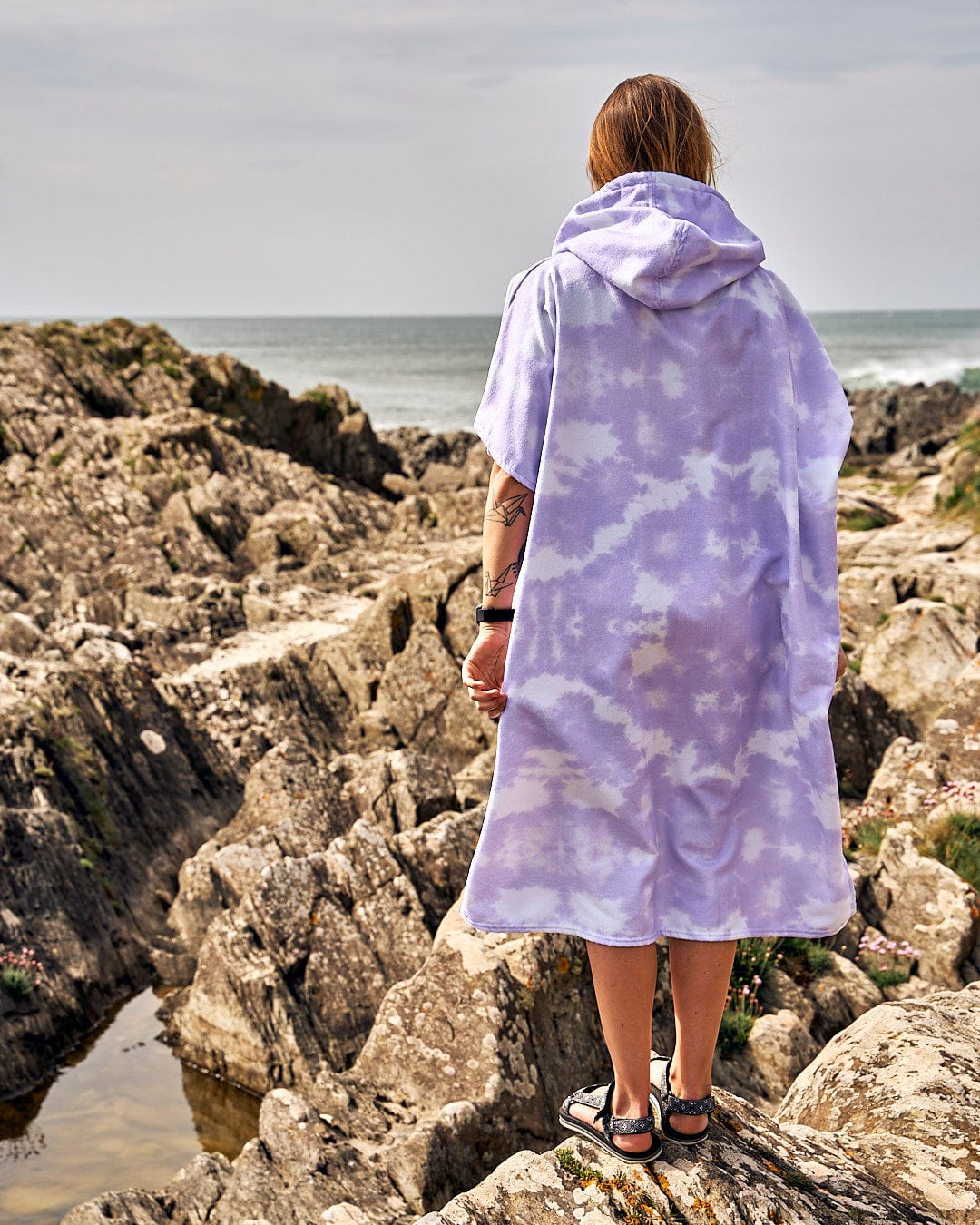 A woman wearing a Saltrock Ubud - Womens Tie Dye Changing Towel - Light Purple poncho standing on a rocky beach.