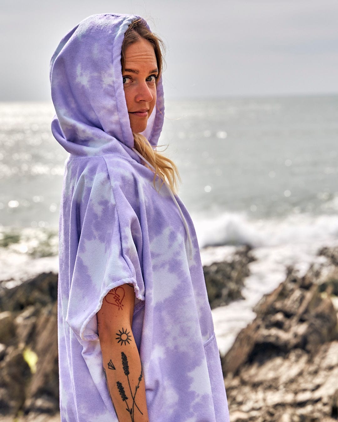 A woman wearing a Saltrock Ubud - Womens Tie Dye Changing Towel - Light Purple hoodie on the beach.
