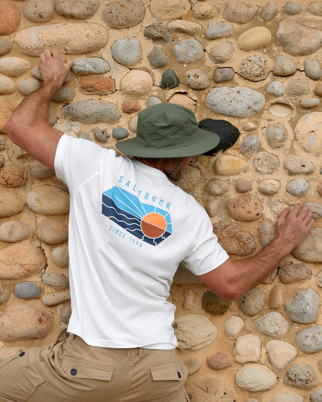 A man in a white t-shirt and tan pants wearing a Saltrock Green Gaitor bucket hat with an adjustable drawstring chin strap is attempting to climb a wall made of rounded stones.