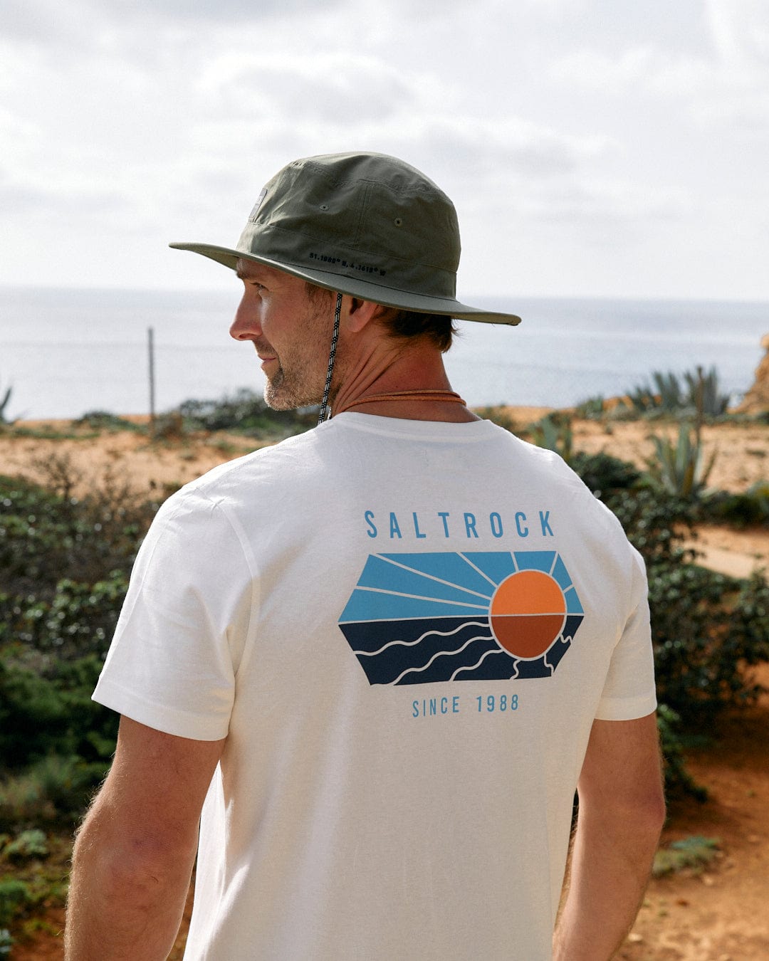 A man wearing a Saltrock Green Gaitor bucket hat and a t-shirt with the "Saltrock since 1988" logo on the back looks out towards a coastal landscape.