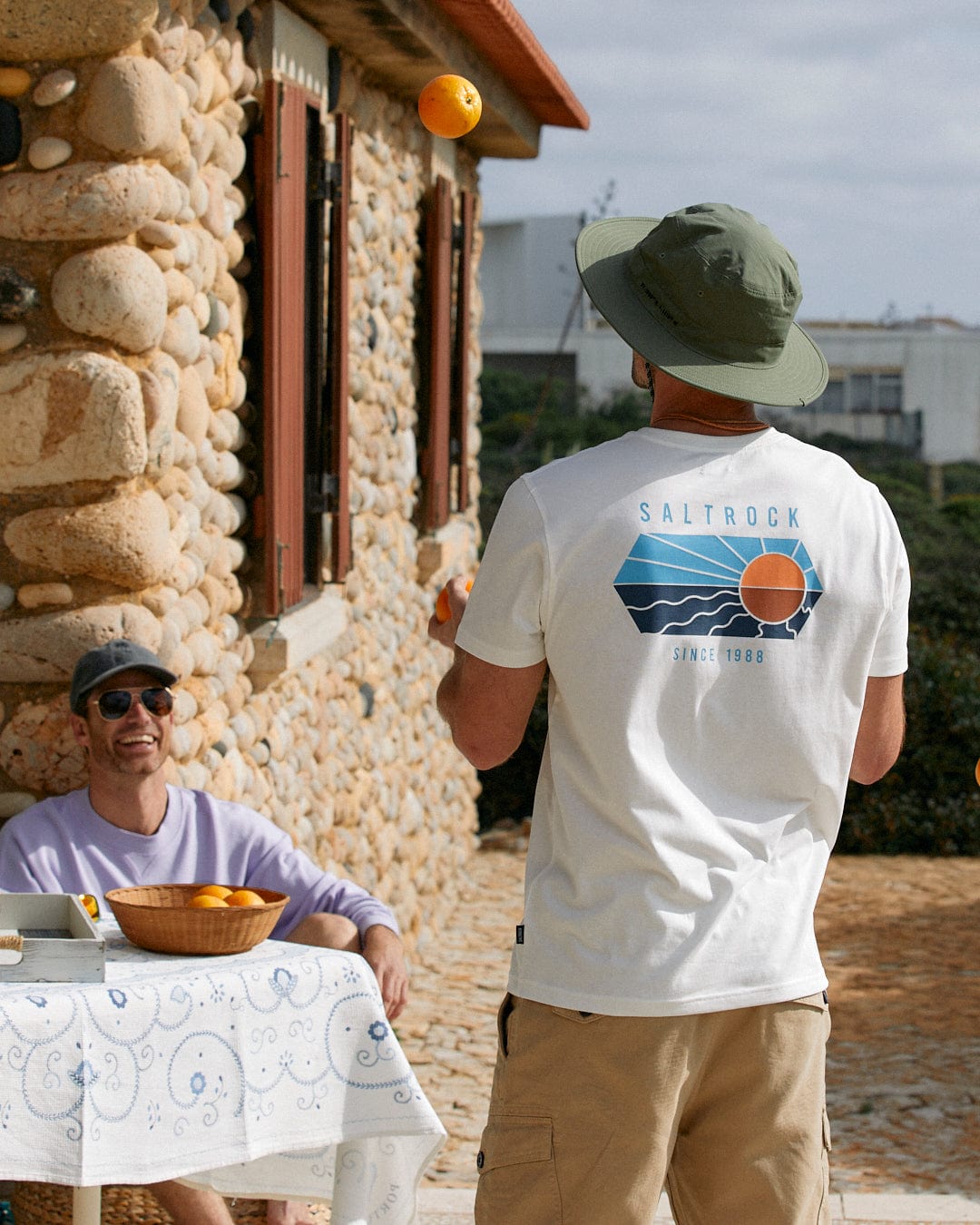 Two men are enjoying an outdoor activity; one is throwing an orange in the air while the other, wearing a Saltrock green Gaitor bucket hat with a water-resistant adjustable drawstring chin strap, watches from a seat at a table.