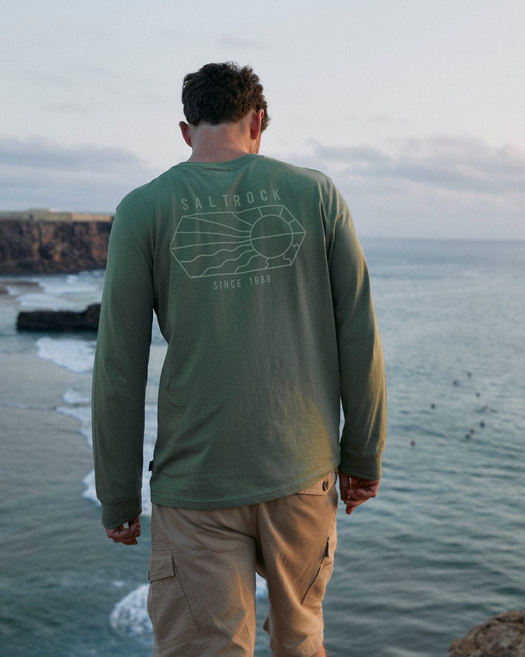 The back of a man standing on a cliff overlooking the ocean, wearing a Saltrock Vantage Outline - Mens Long Sleeve T-Shirt in Green.