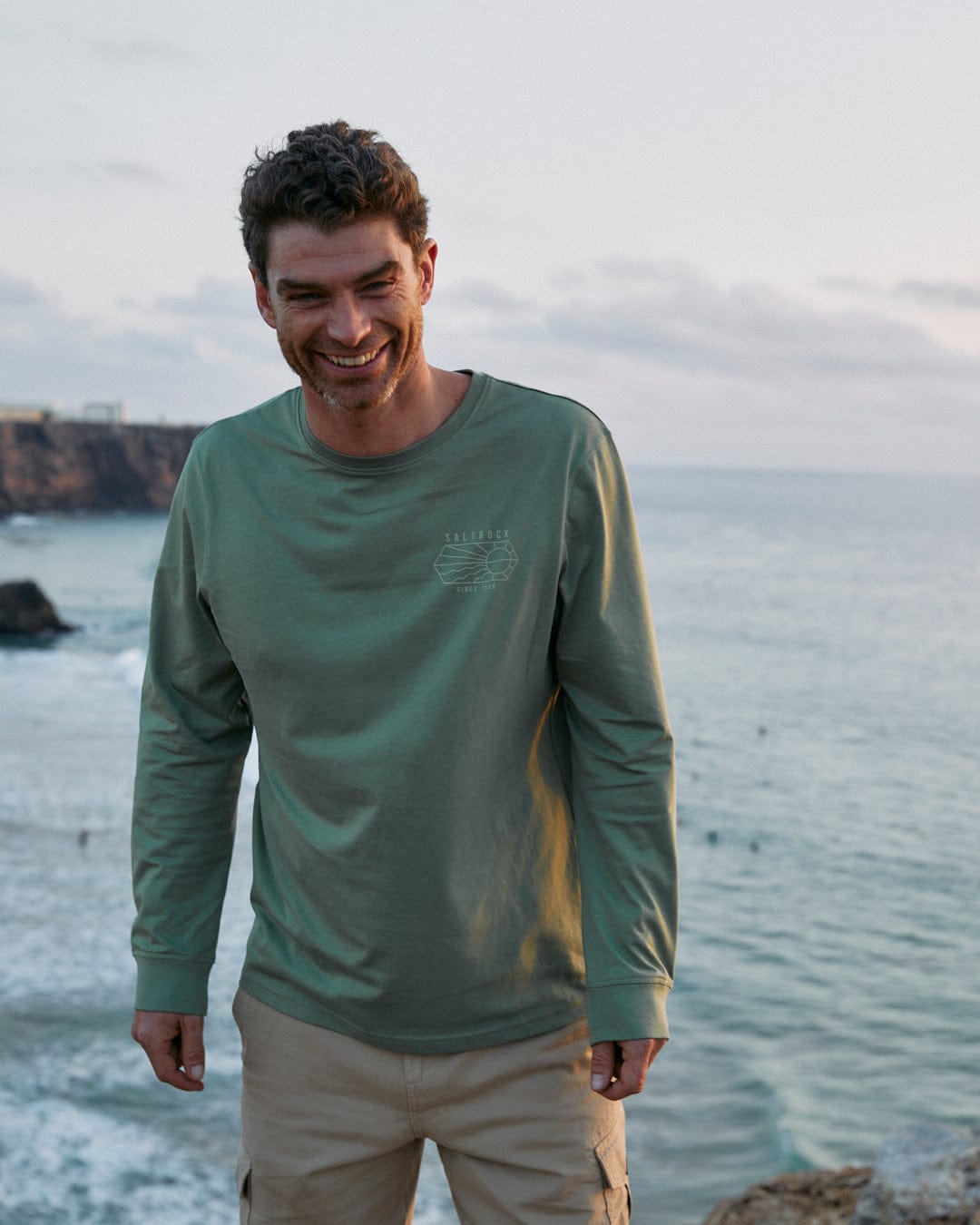 A man standing on a rock near water in a Saltrock Vantage Outline Mens Long Sleeve T-Shirt in Green.