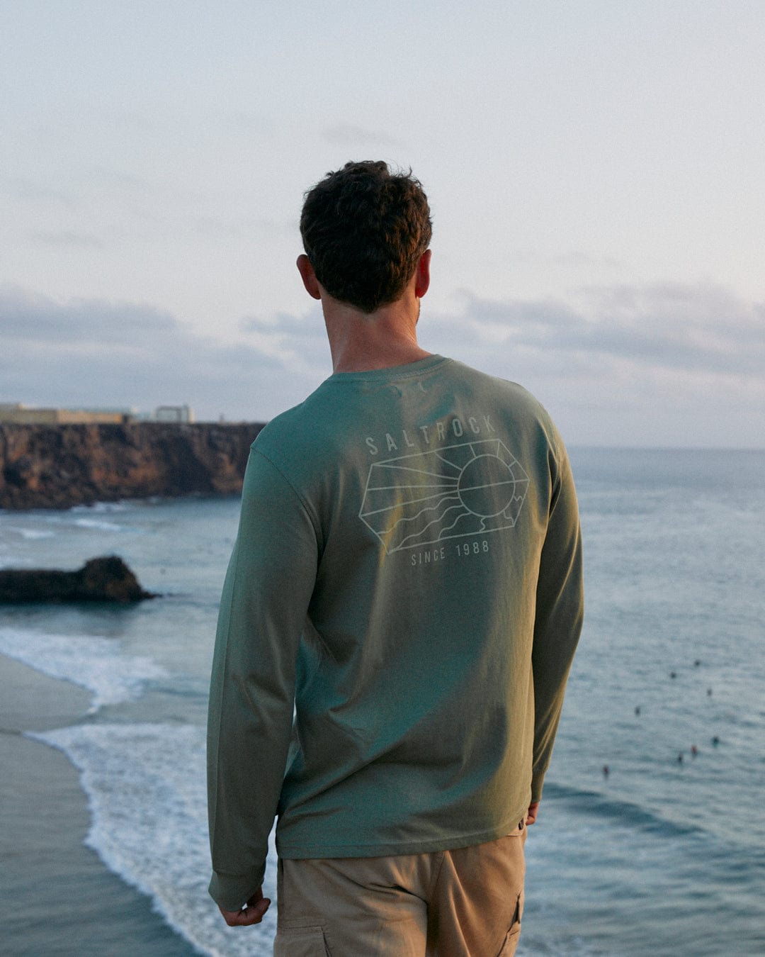 A man is standing on the beach wearing a Saltrock Vantage Outline - Mens Long Sleeve T-Shirt - Green, made of 100% Cotton, looking at the ocean.