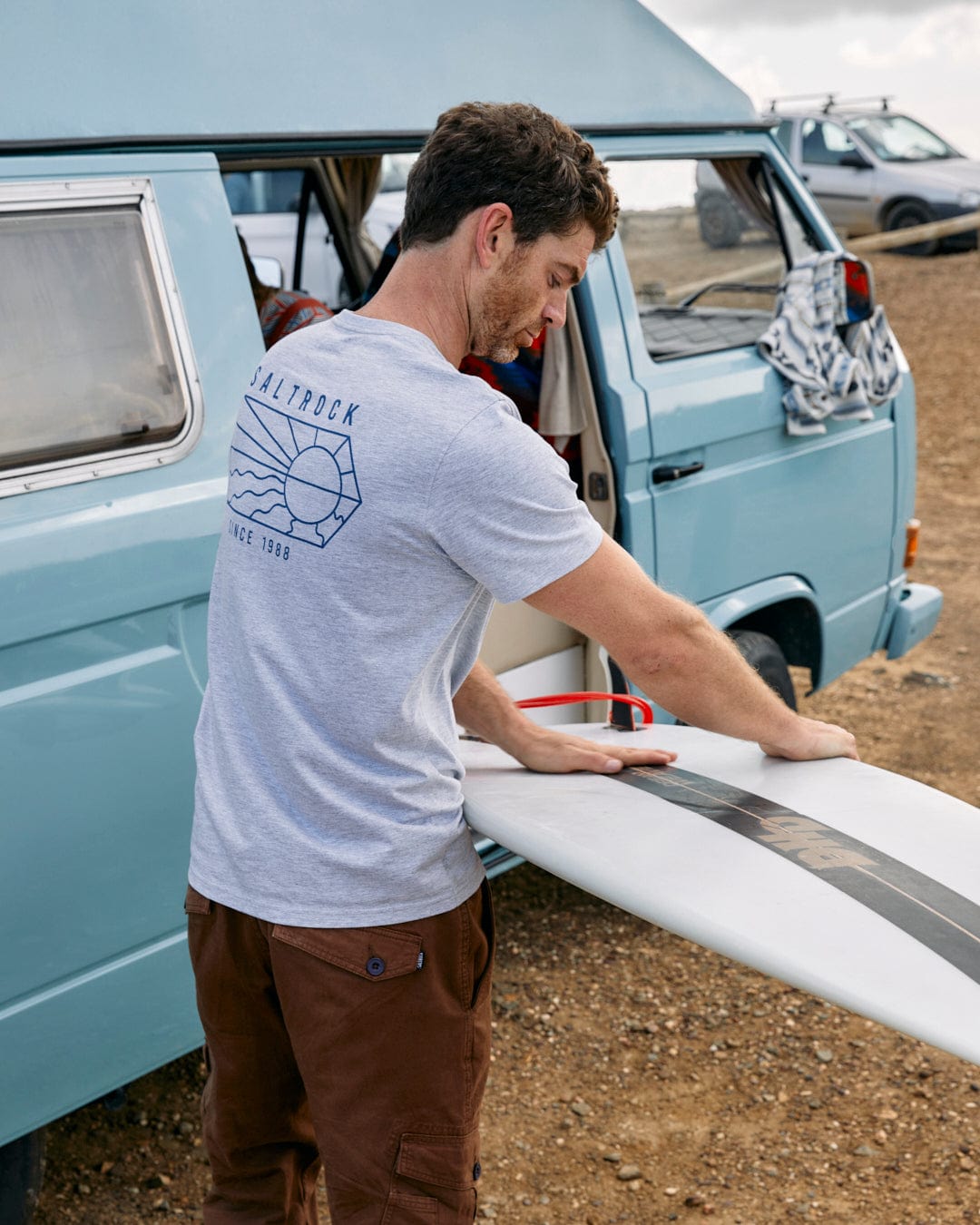 A man with a surfboard wearing a Saltrock Vantage Outline - Mens Short Sleeve T-Shirt in Grey Marl.