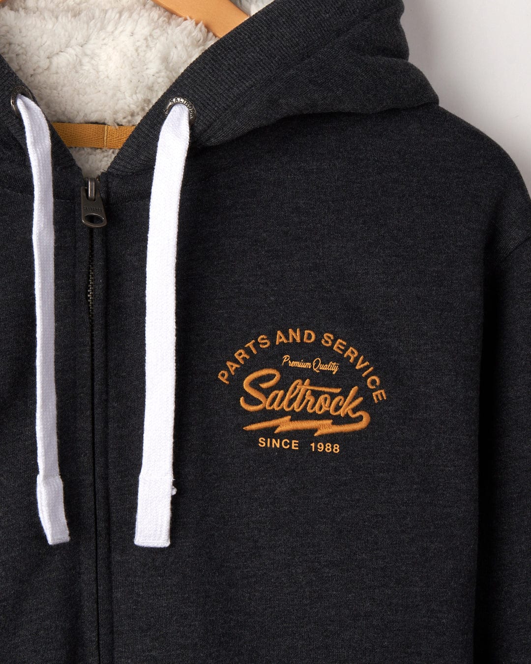 Close-up of a dark gray Vegas Script - Mens Borg Lined Zip Hoodie - Grey with Saltrock branding embroidered in chain stitch on the left side, featuring white drawstrings and a partial view of a Borg-lined hood.