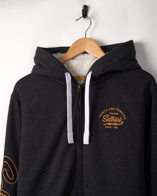 A dark gray Vegas Script - Mens Borg Lined Zip Hoodie - Grey with a white fur lining, featuring an embroidered chain stitch logo "cats and serbs, outbrook, since 1986" on the left chest, hanging on a wooden h
