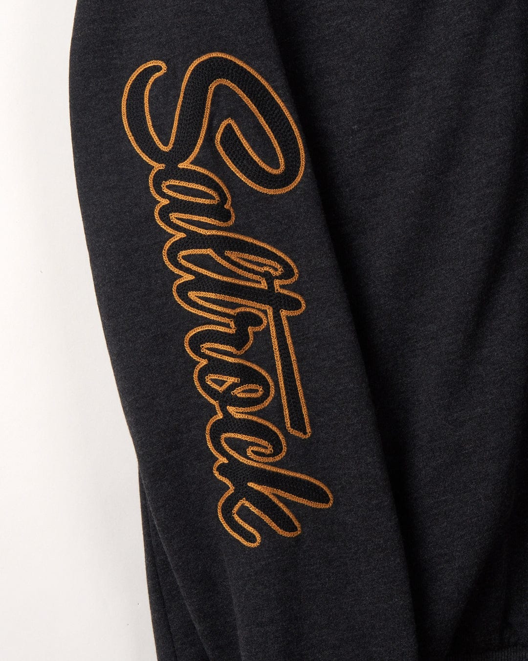 Close-up of a grey Vegas Script Borg Lined Zip Hoodie with a gold "Saltrock" logo embroidered chain stitch in a cursive style down the sleeve.