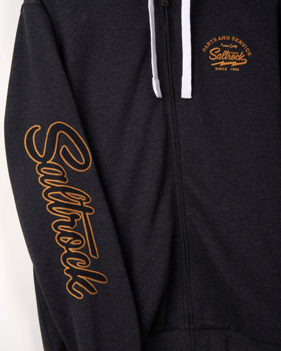 Close-up of a dark gray zip-up hoodie with the embroidered chain stitch logo "Saltrock" in yellow, and text "surf and service since 1988" on the left chest area.