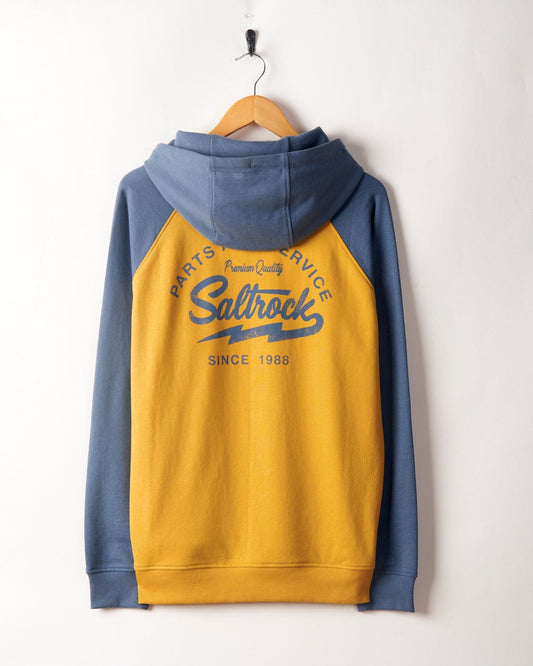 A blue and yellow Saltrock Vegas Script - Mens Pop Raglan Hoodie with raglan sleeves, the logo, and "since 1988" text, hanging on a white wall.