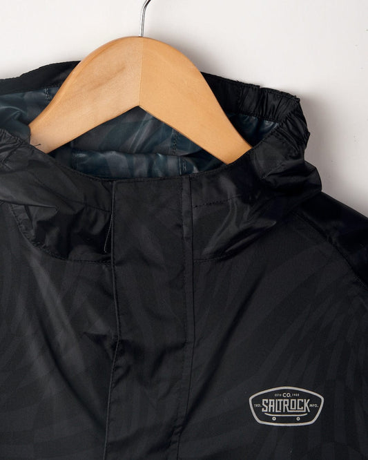 Close-up of a black Saltrock Warped - Kids Waterproof Packable Jacket with a quilted lining on a wooden hanger, featuring a logo patch on the chest that says "Saltrock Co.