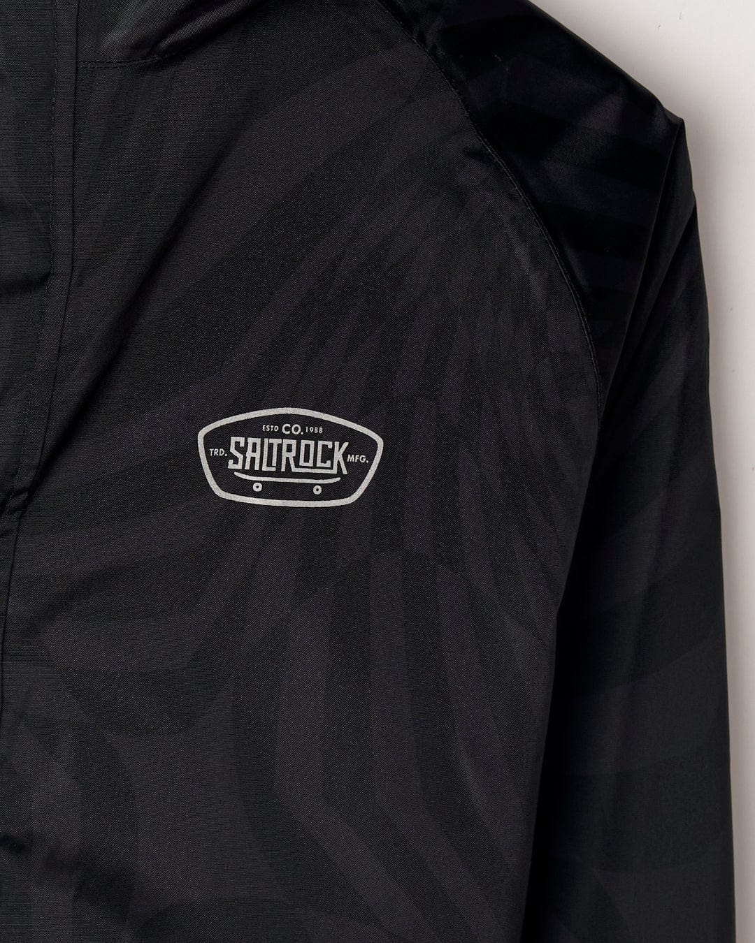Close-up of a black Warped - Kids Waterproof Packable Jacket with a geometric pattern and a white and gray Saltrock logo patch on the shoulder.