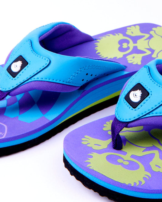 A pair of Saltrock Warp Icon - Kids Flip Flops - Blue with blue/purple toe straps and purple soles, decorated with green monster prints.