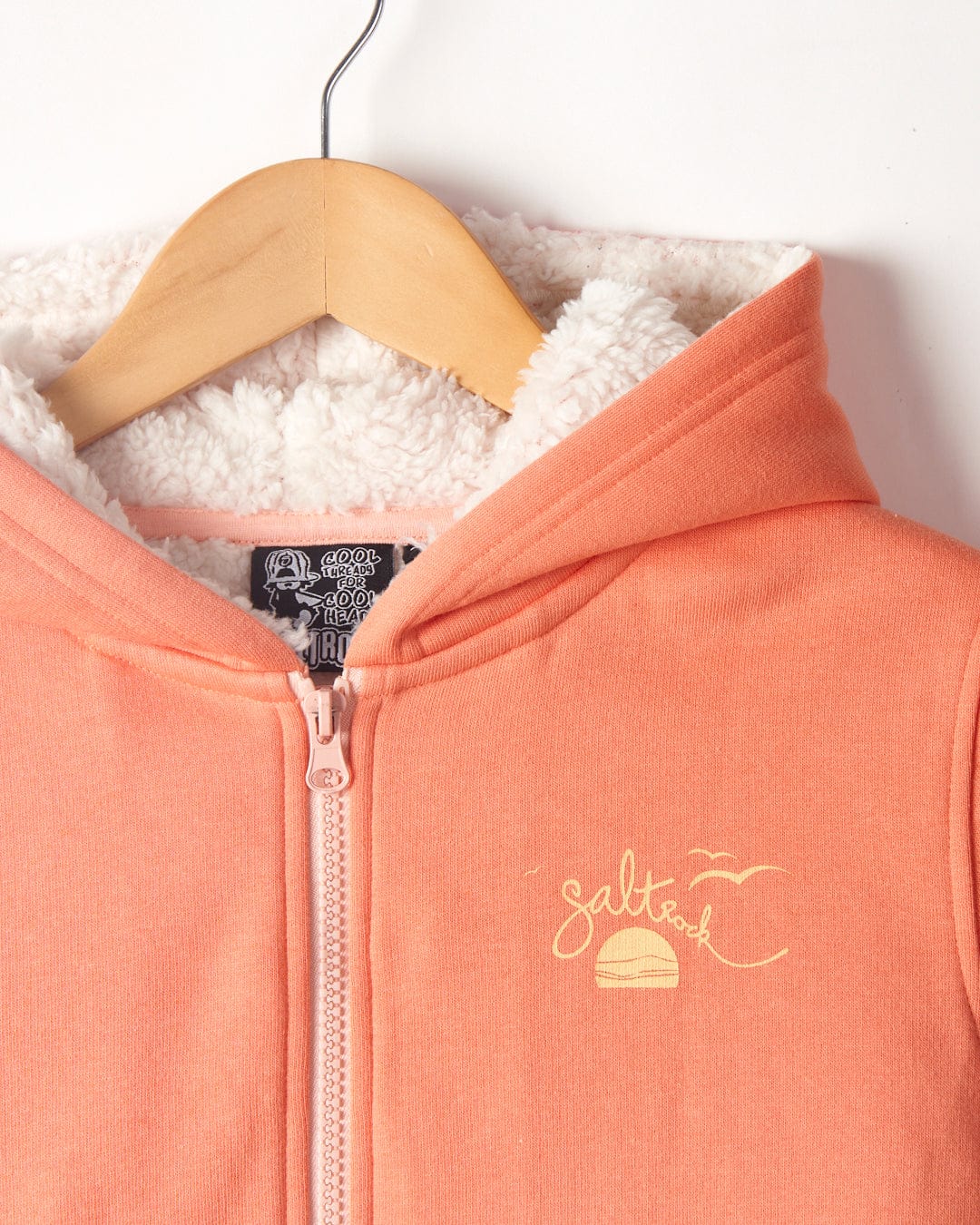 Close-up of a Waveline - Kids Borg Lined Zip Hoodie in coral with a wooden hanger, featuring an embroidered sunset logo and Borg lined inner lining by Saltrock.