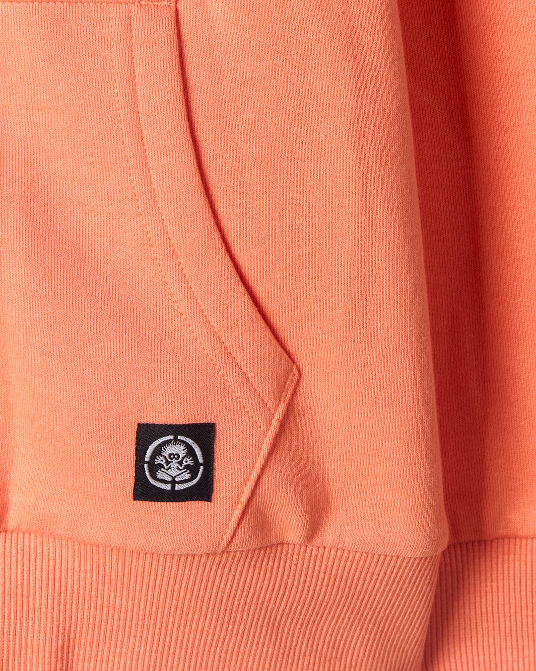 Close-up of a Waveline - Kids Borg Lined Zip Hoodie in Coral by Saltrock, featuring a small black label stitched on with a stylized white lion logo.