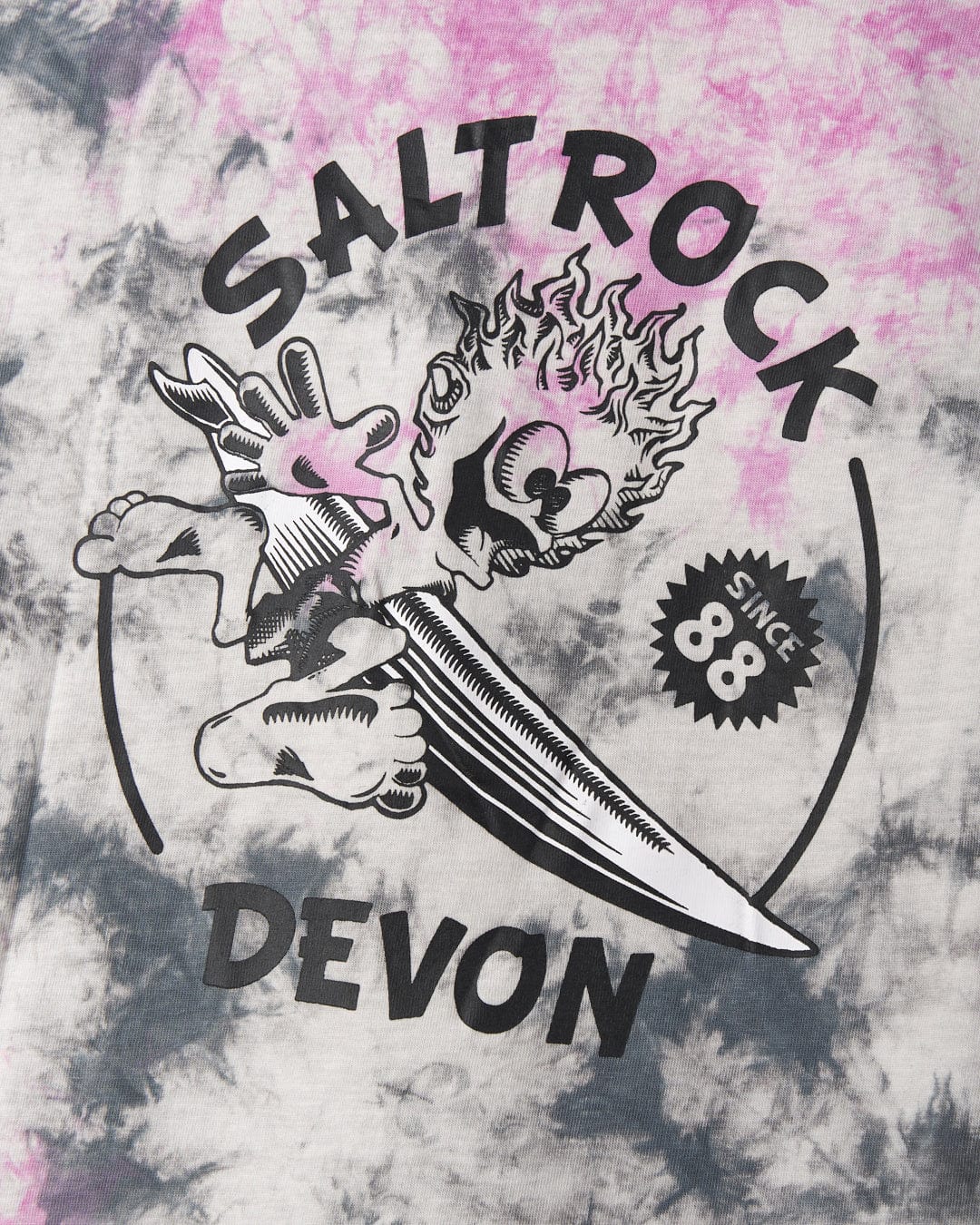 Graphic print on a tie-dye fabric featuring the word "Saltrock" alongside an illustration of a sword and flame, with the text "Devon" and "since 88" below. Made