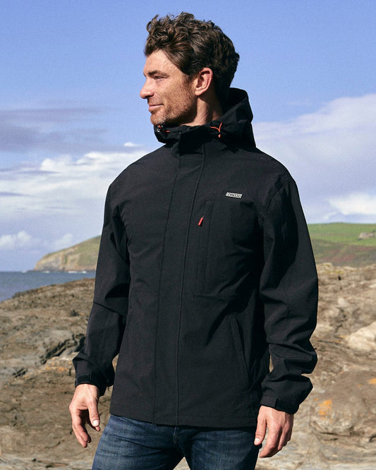 A man, clad in a Saltrock Whistler - Mens Hooded Jacket - Black with mesh lining, stands on a rocky beach.
