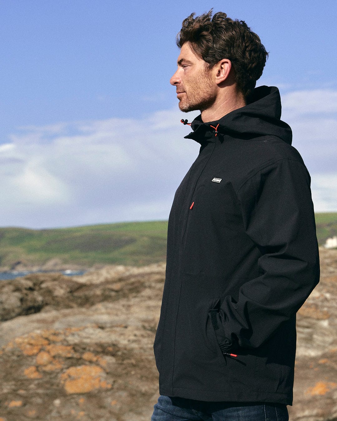 A man, wearing a Saltrock Whistler - Mens Hooded Jacket - Black with zip pockets, is standing on a rock looking at the ocean.