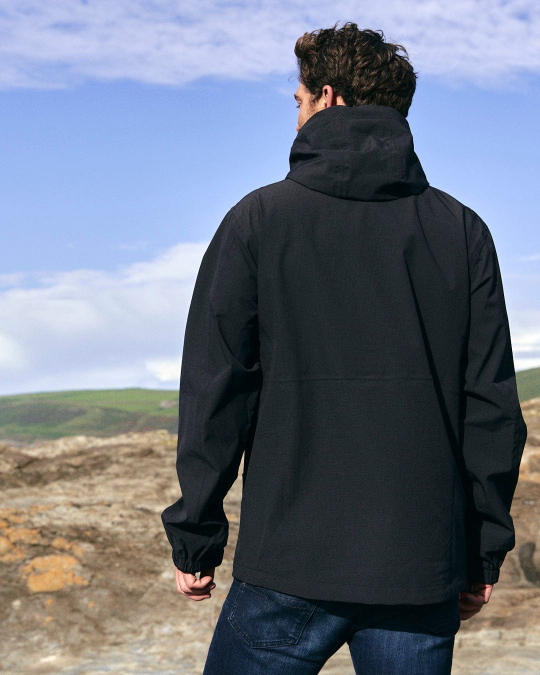 A man in a Saltrock Whistler - Mens Hooded Jacket - Black standing on a rocky beach.