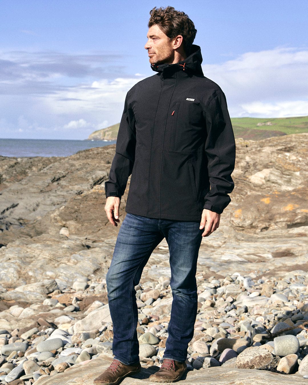 A man standing on a rocky beach wearing a Saltrock Whistler - Mens Hooded Jacket - Black.