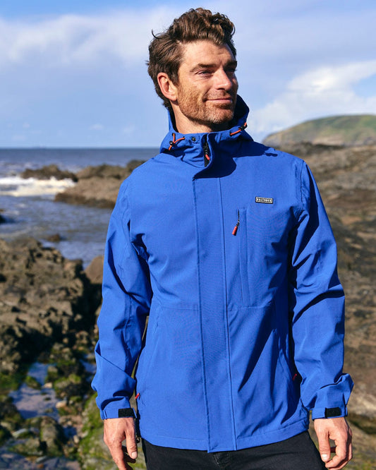 A man in a Saltrock Whistler Mens Hooded Jacket in Blue standing on rocks near the ocean.